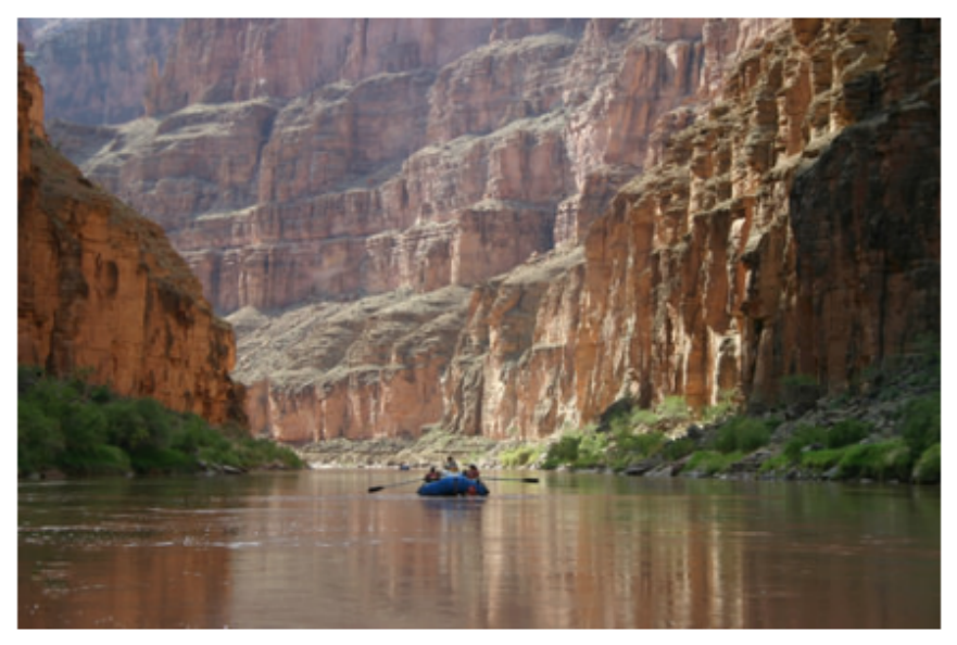 Photograph of a river with a small raft, flowing between tall, layered canyon walls.